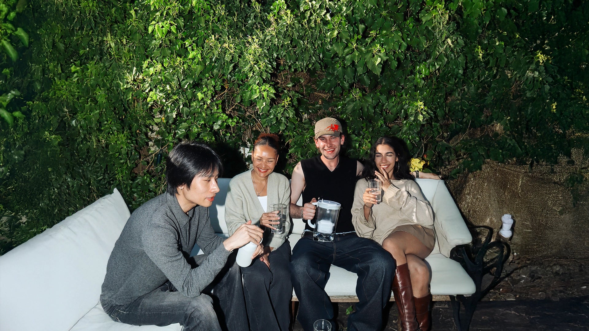 Group of four people sitting outdoors on a white sofa surrounded by green foliage, holding glasses of water, a BRITA water filter jug, and a BRITA water filter bottle during a social gathering.