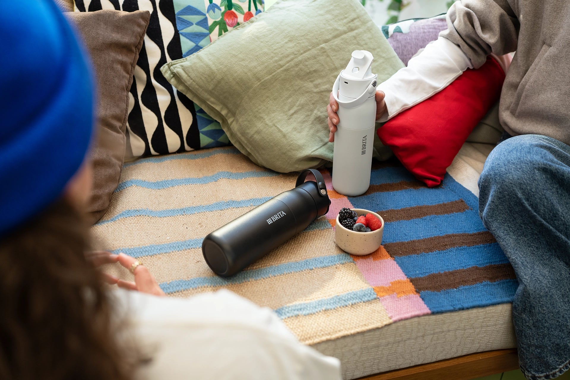 Two people sitting on a colourful couch with black and white BRITA LARQ Bottles, next to a bowl of fresh berries and decorative cushions.