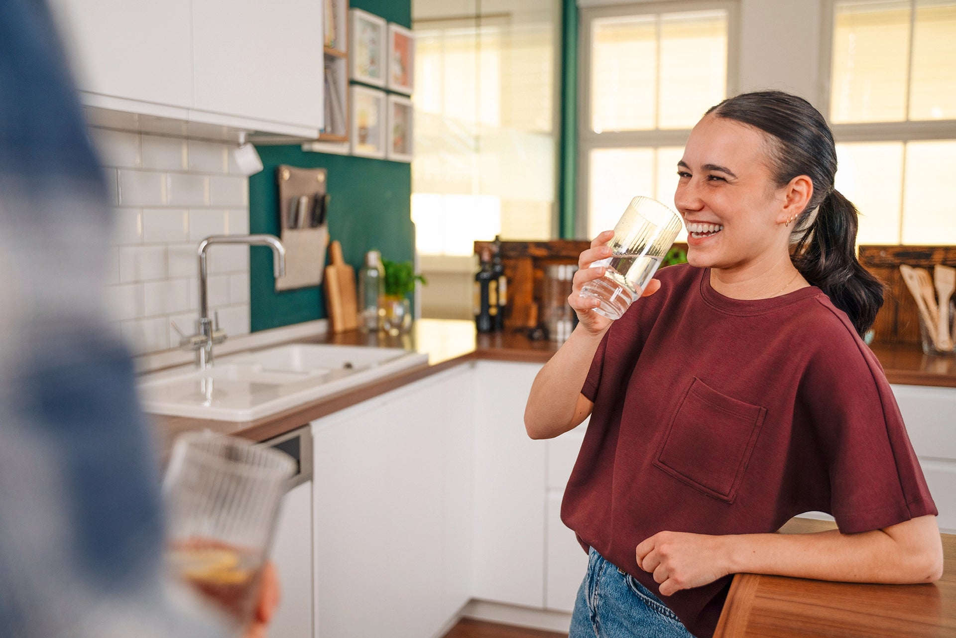 Smiling woman holding a glass of water in a kitchen, with another person in the foreground.