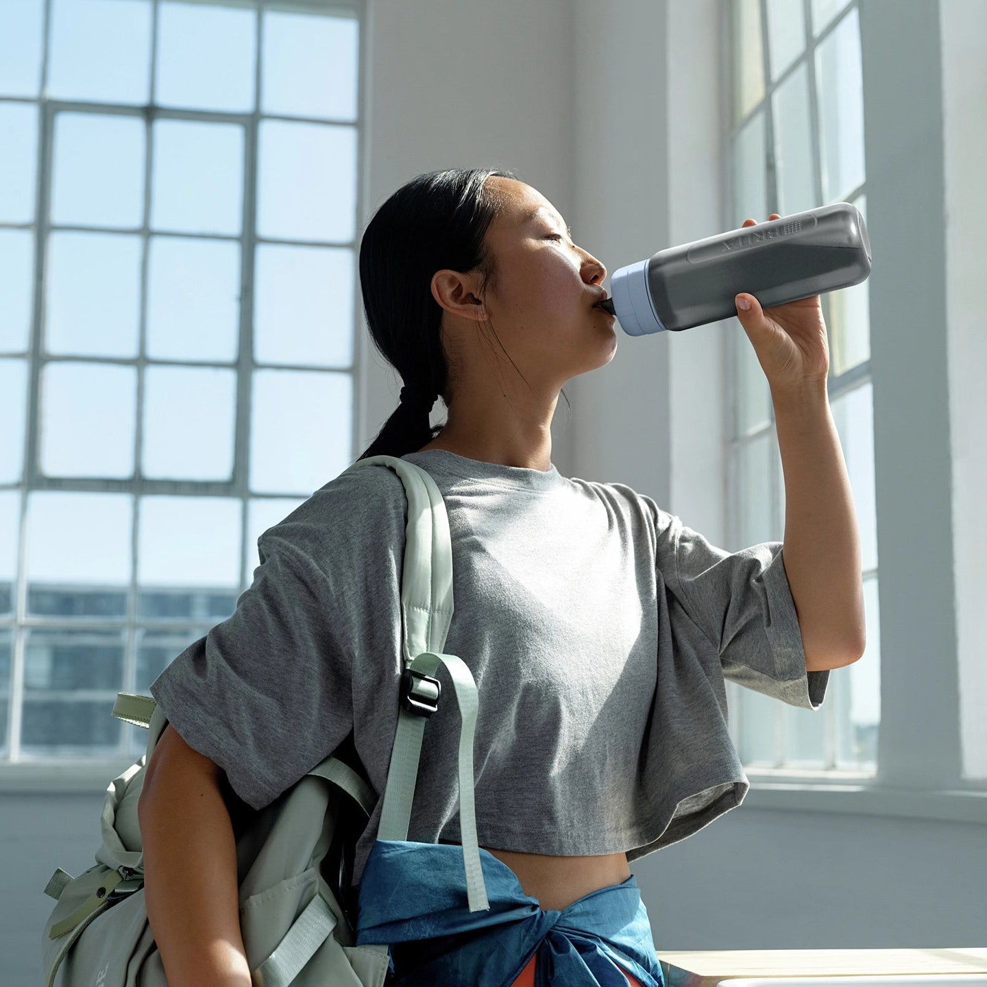 Una mujer bebiendo de una jarra de agua BRITA filtro Modelo «Bottle» en una habitación luminosa con ventanas grandes.