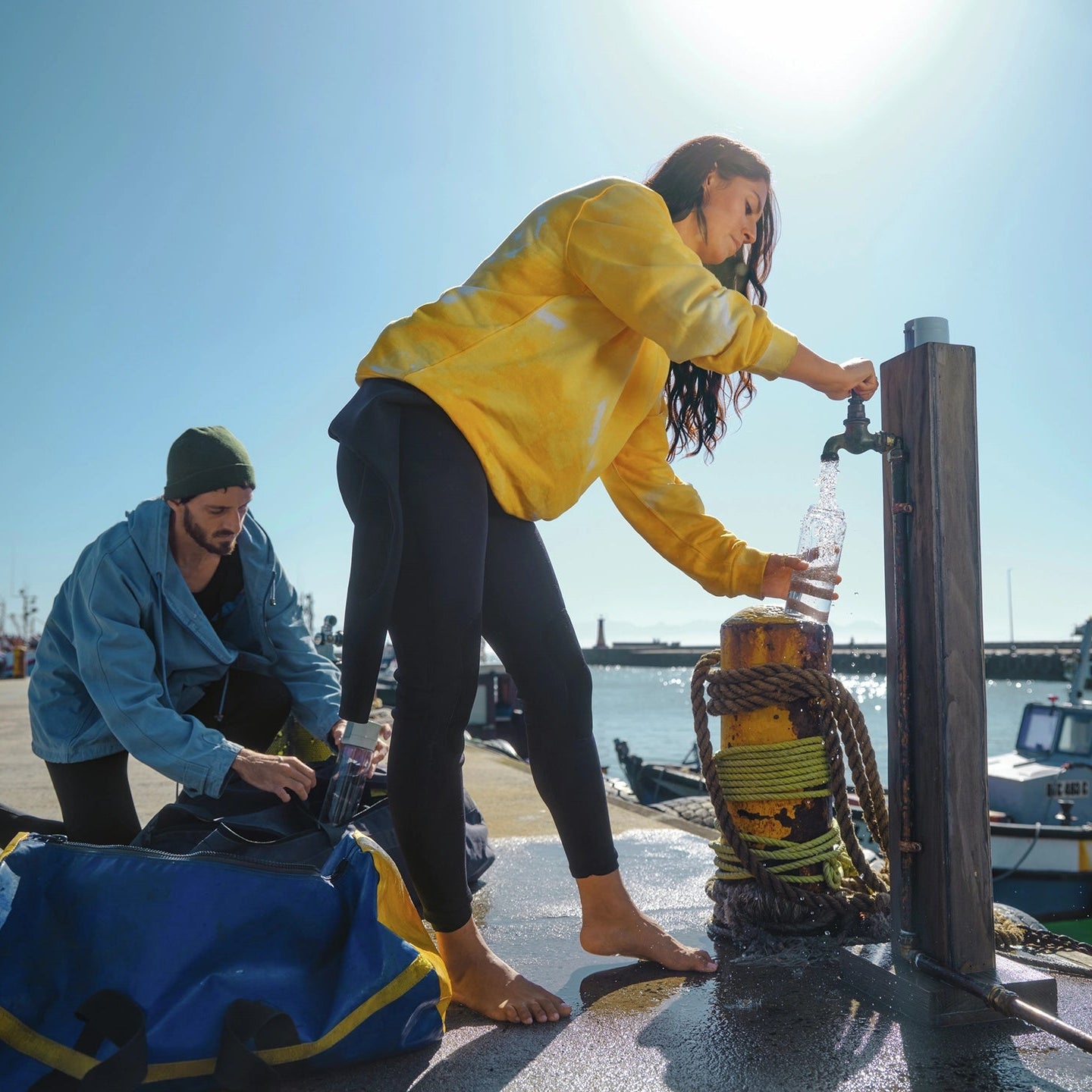Hay dos personas en un muelle junto a un cuerpo de agua; una de ellas está usando un grifo para llenar una jarra BRITA. filtro Botella modelo Vital.
