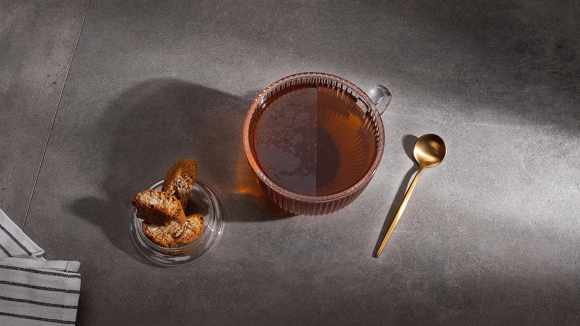 A clear ribbed glass mug filled with tea sits on a dark gray surface, alongside a small glass dish of cookies and a gold spoon.
