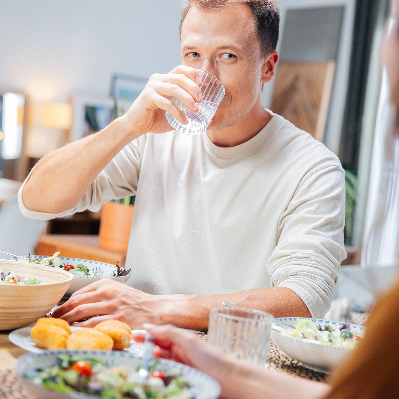 Un hombre bebiendo agua en una mesa de comedor con comida y otra persona que se ve parcialmente